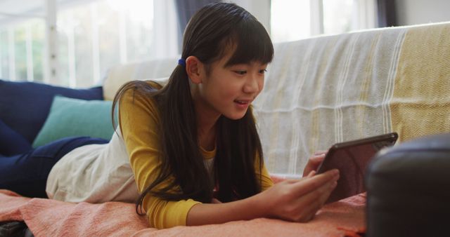 Young Asian girl lay on couch using tablet at home. Ideal for educational, family, and domestic lifestyle topics. Represents comfort, technology use amongst children, and modern family living.