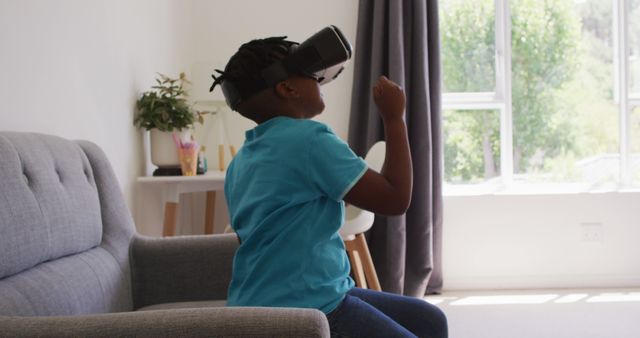 Image of a young African American boy sitting on a sofa in a bright living room, wearing a VR headset and engaging with virtual reality. Great for use in technology articles, educational materials, creative campaigns, or advertisements related to modern gaming, interactive learning, and children's engagement with tech.