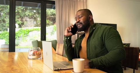 African American man working from home talking on phone while using laptop at dining table