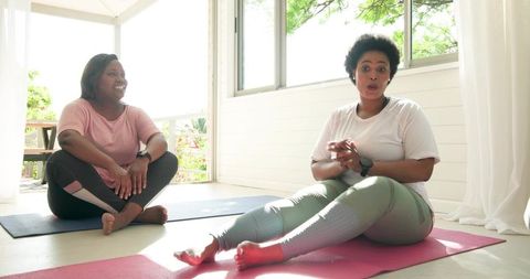 Two women exercising and chatting on sunlit porch mats, stretching and laughing together