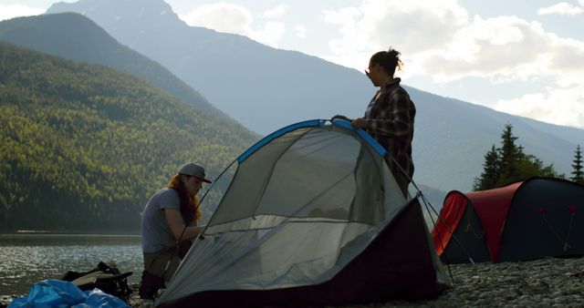 Two friends setting up tent during camping trip by lake surrounded by scenic mountains. Useful for themes related to adventure, outdoor activities, friendships, camping, nature trips, teamwork, and travel promotions.