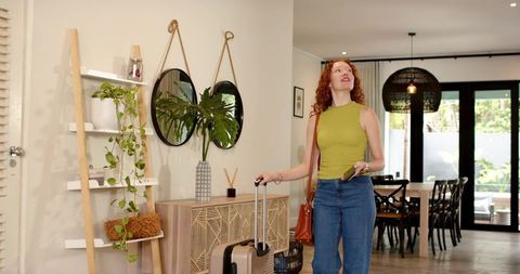 Redhead woman standing in sunlit entryway with wheeled suitcase and smartphone