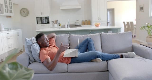 Man lounging on a grey sofa while engaging in a video call on a laptop in a modern, cozy living room. Brightly lit space features stylish kitchen setup in background. Ideal for depicting remote communication, modern lifestyle, comfort at home. Suitable for articles on home technology, lifestyle blogs, and remote work scenarios.