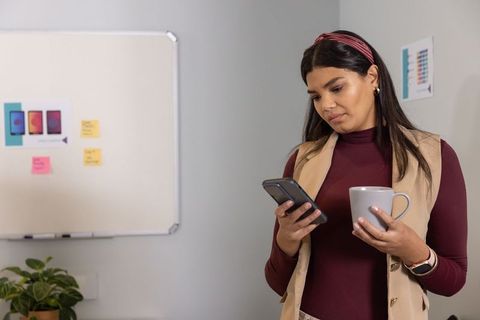 Mid-adult woman checking phone while holding coffee and reviewing whiteboard in office