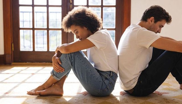 Couple sitting back-to-back on rug in sunlit room conveying emotional distance