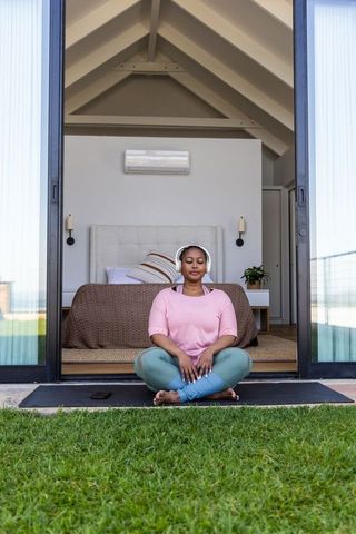 African american woman meditating at bedroom doorway wearing headphones, phone nearby
