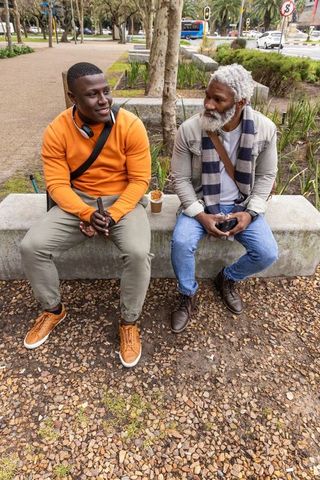 African american men sitting on bench in urban park, senior and younger talking with smartphones