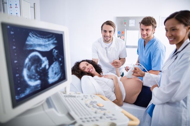 Doctors performing an ultrasound scan on a pregnant woman in a hospital. The woman is lying on a bed, smiling, while the medical professionals are focused on the ultrasound monitor. This image can be used for articles on prenatal care, medical examinations, healthcare services, maternity care, and teamwork in medical settings.