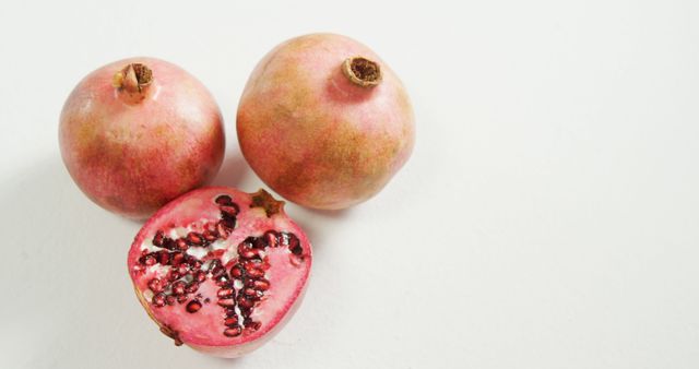 This image showing one cut pomegranate with red seeds exposed and two whole pomegranates on a white background is perfect for health and wellness articles, blogs promoting healthy eating, fruit advertisements, and dietary advice content. The vibrant color contrast between the red pomegranate seeds and the white background enhances visual appeal, making it ideal for food-related publications.