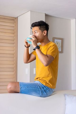 Asian man sipping coffee on couch back in modern living room wearing mustard t-shirt