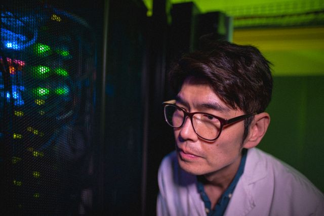Asian male technician is closely examining servers in a data center. He is wearing glasses and a white lab coat, indicating a professional setting. The image highlights technology, cloud computing, and networking themes. Ideal for use in articles, blogs, or advertisements related to IT, data management, server maintenance, and technological advancements.