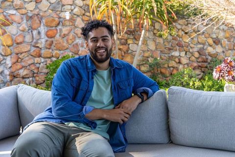 Smiling man relaxing on patio sofa in denim shirt and khaki pants with garden backdrop