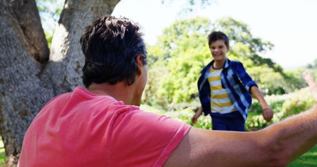 Father extending arms to his son in a park setting. Boy happily running towards father with greenery and trees in the background. Ideal for promoting family activities, parenting, and outdoor play.