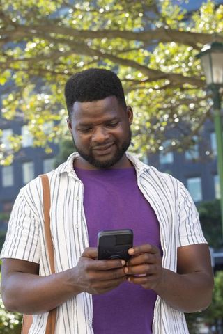 African american man checking smartphone in sunlit urban plaza with shoulder bag