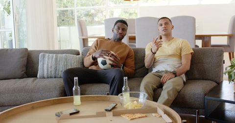 Diverse friends watching soccer on couch holding ball pointing at tv snacks drinks relaxed weekend