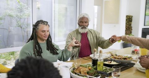 Multi-generational family holding hands and praying around sunlit holiday dinner table