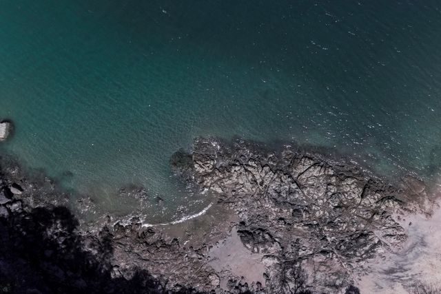 Aerial view capturing the serene and tranquil coastal scene with rocky shorelines meeting the calm sea. Ideal for incorporating into projects focused on nature, coastal conservation, environmental themes, or travel promotions highlighting serene coastal destinations.