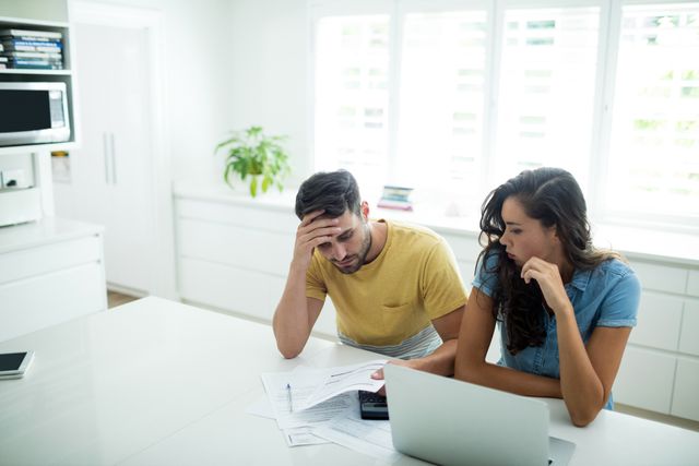 Young couple sitting in modern kitchen, looking stressed while calculating invoices on laptop. Ideal for content related to financial planning, budgeting, family expenses, and domestic life. Can be used in articles, blogs, and advertisements focusing on financial advice, home budgeting, and stress management.