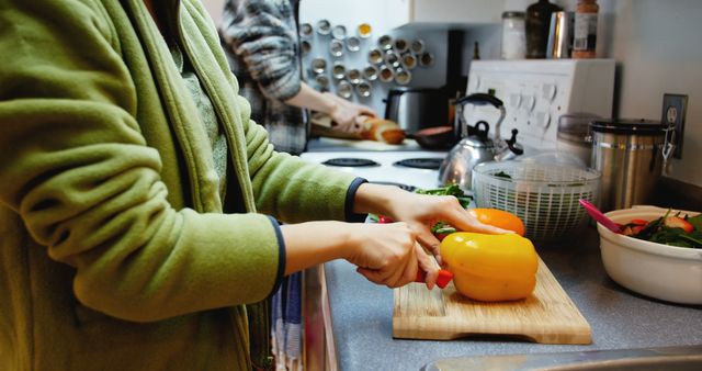 Individuals are preparing fresh vegetables in a modern kitchen, focusing on cutting yellow and orange peppers. Ideal for use in cooking blogs, healthy lifestyle articles, meal preparation guides, and advertisements for kitchen appliances or fresh produce.