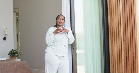 Middle-aged african american woman relaxing in pajamas on sunlit bedroom balcony with mug