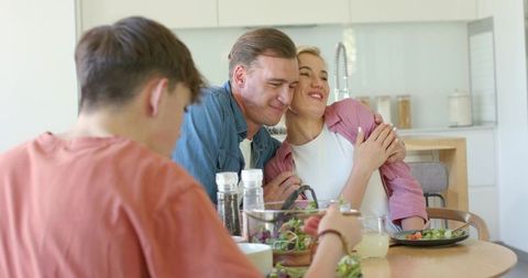 Parents embracing at kitchen table while teen serving salad, warm family moment
