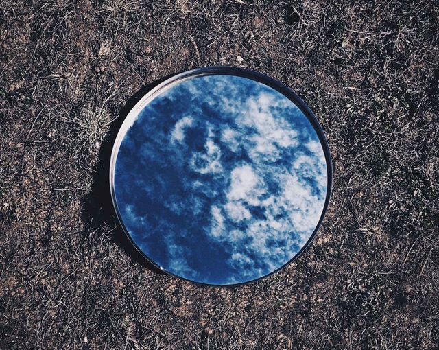 Circular mirror placed on earthy ground reflecting blue sky and white clouds above. Artistic and abstract photograph ideal for design projects, nature-themed presentations, and optical illusion galleries. Useful as a visual metaphor for introspection, self-reflection, and the connection between earth and sky.