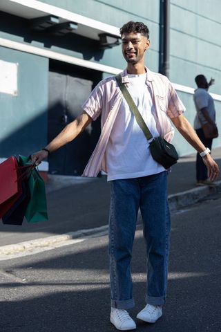 Young man carrying colorful shopping bags on sunny urban street, wearing crossbody bag