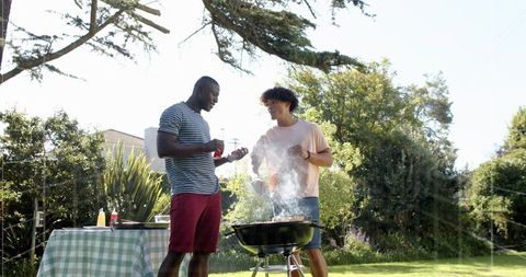 Two friends grilling in sunny backyard with charcoal grill and checkered picnic table