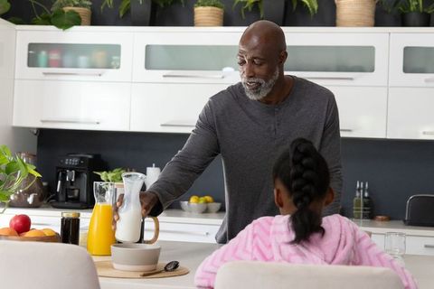 African american dad pouring milk into cereal for daughter at bright modern kitchen island