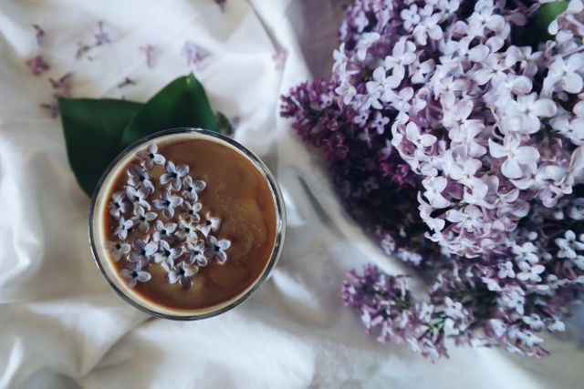 Top-down view of iced coffee topped with beautiful lilac blossoms, placed on white cloth beside lilac flowers. Ideal for use in food blogs, coffee shop promotions, floral decoration articles, and lifestyle magazines.