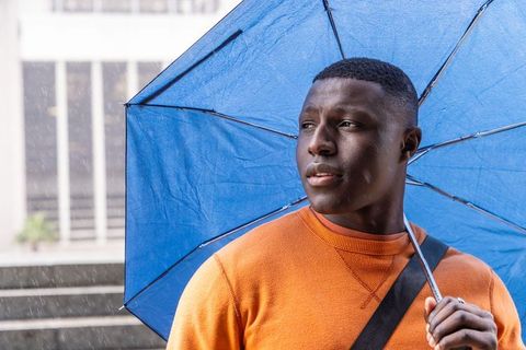 Thoughtful african american man holding blue umbrella on rainy urban steps