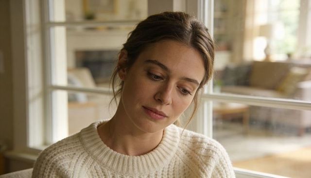 Contemplative woman wearing cream knit sweater reflecting by window in cozy living room