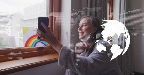 Woman taking selfie at window with rainbow drawing, mask lowered and digital globe overlay