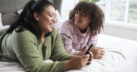 Two friends laughing and sharing smartphone while lounging on bed in bright cozy bedroom