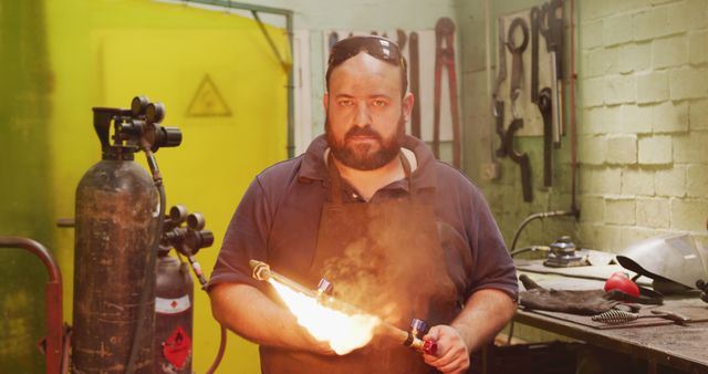 Bearded male metalworker using an oxy-acetylene torch in an industrial workshop. He is wearing safety gear and surrounded by various tools and equipment. This image is perfect for illustrating skilled craftsmanship, metalworking, or industrial labor. Great for use in websites, brochures, or articles related to manufacturing, engineering, or vocational training.