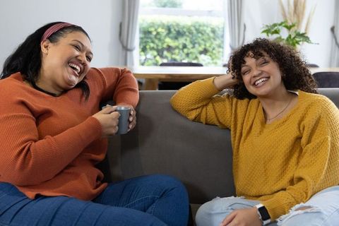 Two african american women laughing and chatting on couch sharing cozy coffee moment