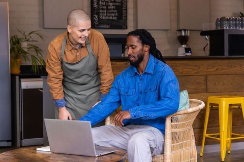 Barista helping african american customer using laptop in cozy coffeehouse with wicker seating