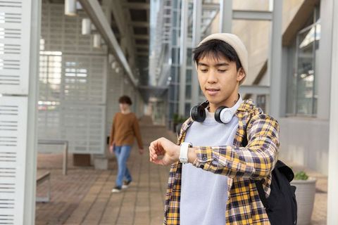 Asian student checking smartwatch on campus walkway wearing headphones, beanie and backpack