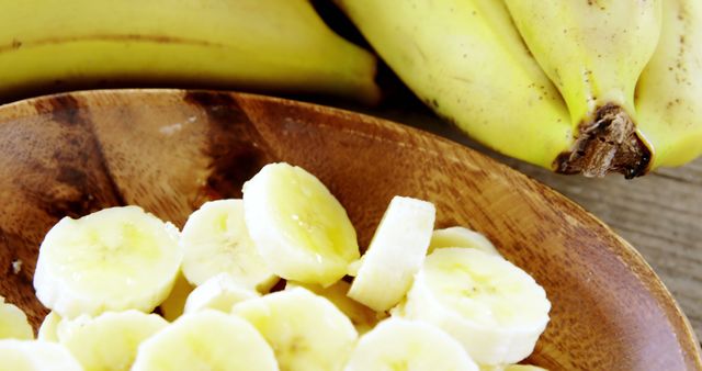 A wooden bowl contains sliced bananas, with a bunch of whole bananas in the background, with copy space. Fresh bananas, both whole and sliced, are presented, suggesting a healthy snack or ingredient preparation.