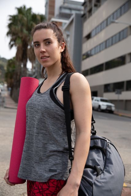 A fit Caucasian woman walking in the street on her way to fitness training, carrying a sports bag and a yoga mat