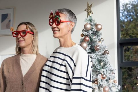 Mature women wearing novelty glasses beside flocked christmas tree, smiling together during holidays