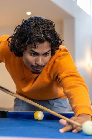 Indian man in twenties leaning over blue pool table aiming cue with focused concentration