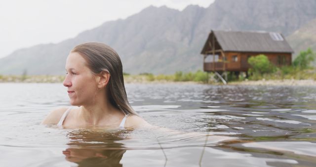 Woman enjoying swim in pristine mountain lake. Rustic cabin on shore and mist-shrouded peaks in background. Perfect for themes of adventure, nature retreats, outdoor leisure, tranquility, and serene getaways.