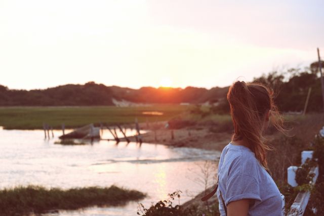 Woman standing by a riverside during a beautiful sunset, gazing at the horizon. Ideal for concepts related to relaxation, nature, summer travel, peaceful getaways, and enjoying the outdoors.