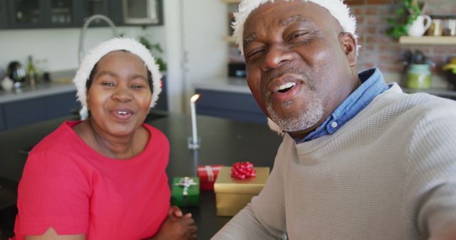 Senior couple wearing Christmas hats and smiling in kitchen, with wrapped gifts in background. Suitable for holiday promotions, advertisements focused on family and joy, and greeting cards.