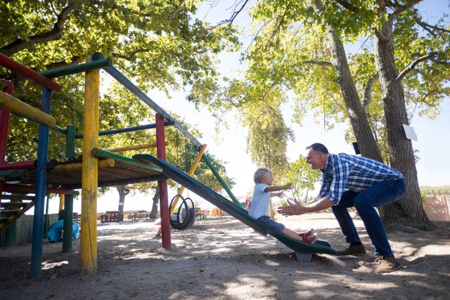 Father encouraging his young son as he slides down a playground slide. The scene is set in a park with trees providing shade. This image is ideal for use in parenting blogs, family activity promotions, and advertisements focusing on family bonding and outdoor fun.