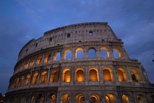 Colosseum in Rome glowing with warm lights against dusk sky. Ideal for travel brochures, historical education, and cultural presentations. Perfect for illustrating ancient Roman architecture and tourism promotions.