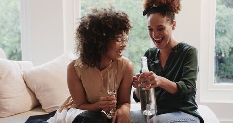 Laughing Diverse Female Friends Pouring Sparkling Wine on Bright Sofa for Casual Celebration