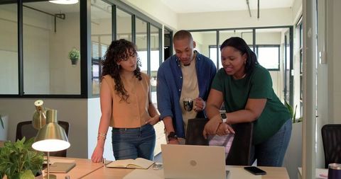 Diverse team collaborating around laptop in modern open-plan office, pointing and smiling