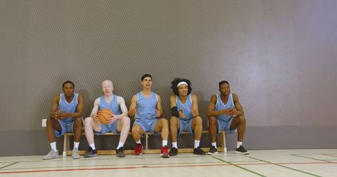 Diverse basketball team sitting on bench in blue uniforms with ball